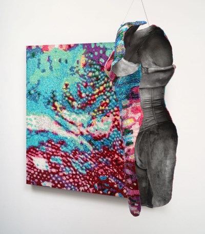 A sculpture hangs in front of a painting. One side of the sculpture and the painting have a similar camouflaged, bejewled pattern. The back of the sculpture has a cast of a body in a corset.  