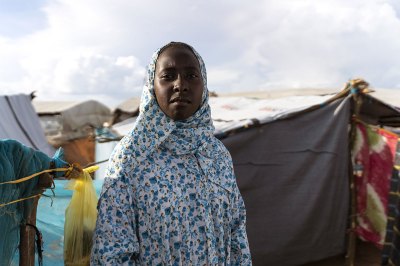 A woman in matching dress and headscarf staring at the camera in blue and white floral prints. She stood in a refugee camp. 