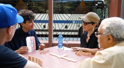 A group of four people sit at a table playing dominos as they talk.
