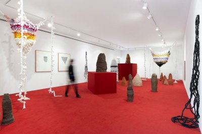 The gallery exhibition has landscapes, sculptures on the floor and pedestals, as well as fiber works hanging from the ceiling and walls. A blurred man walking in the center. 