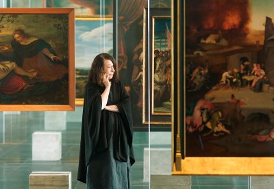 A woman stands in a glass easel designed by Masp by the iconic Lisa Bo Bardi, with an old master painting hanging on it. 