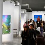 A crowded aisle, at the art fair, all kinds of people searching for paintings at different stalls.