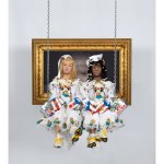 Sculptures of two twin sisters - one with light skin and blond hair, the other with black skin and black hair - on a white communion dress, sneakers and boxing gloves on the swing, decorated with Puerto Rican flags.