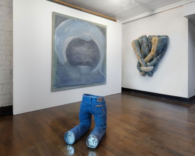 A pair of sculpted legs wearing jeans. Behind this work is a large abstract painting, a wall sculpture composed of a pair of jeans that have been woven together.