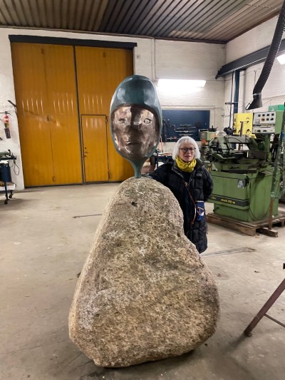 A woman standing next to the head of a carved woman, planted in a piece of granite.