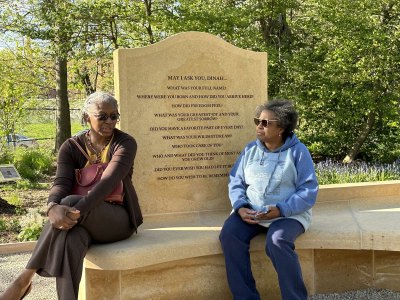 Two black women sat on a bench for public artworks with a slate behind them with various questions engraved on them. 