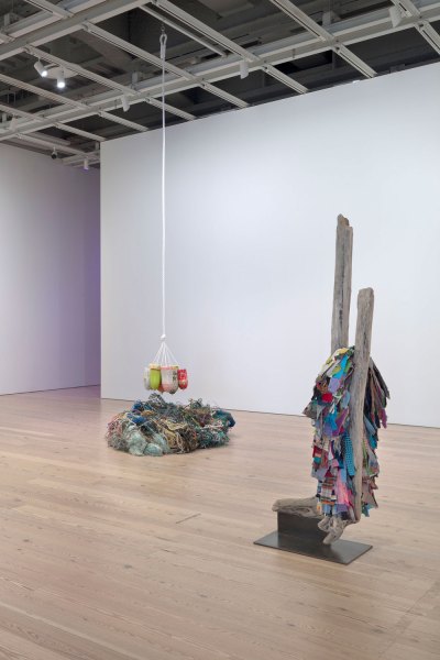 View of two sculptures in the museum. In the foreground, the found clothes are placed between the two branches of the found driftwood. Behind are a bunch of fishing nets found with floats hanging on them. 