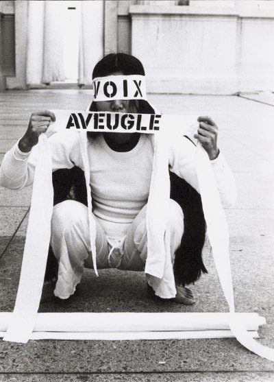 A woman in white dress squatted on the sidewalk. Her eyes and mouth were covered with a white cloth that said 