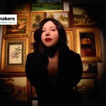 A woman with a black blazer on looks at the camera in front of many artworks in frames.