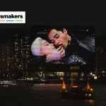 A several-story screen in an urban landscape shows two figures dressed in black with one placing a strawberry into the other's mouth.