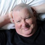 A man lies on the bed with his hands behind his back.