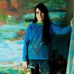 A white woman in a blue sweater playing paint puts one hand on an abstract canvas with the other behind her.