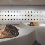 In a large gallery, two boulders are placed on thick white foam rectangles. Along the back wall are three rows of works on paper, all of which include a black circle with a yellow outer ring.