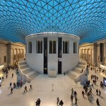 In the color photo, a tan cylindrical structure is seen under the blue curved canopy. In this covered space, many people roam. Neoclassical buildings are at the back.
