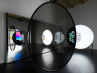 View of the gallery exhibition, showing the various circles, with video projected on it. A mirror can be seen in the background. 