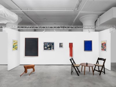 A table and chair sit in front of several pieces hanging on a white wall.