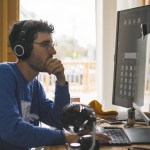 A man wearing headphones and blue sweater sitting on a computer.