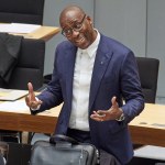 A black man in a suit stood in front of a low table with a briefcase on it. He raised his hands and leaned his head aside.