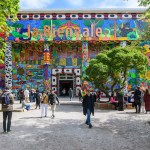 People walk into and out of a treed courtyard in front of a building painted on a colorful pillar. On the roof is the term 