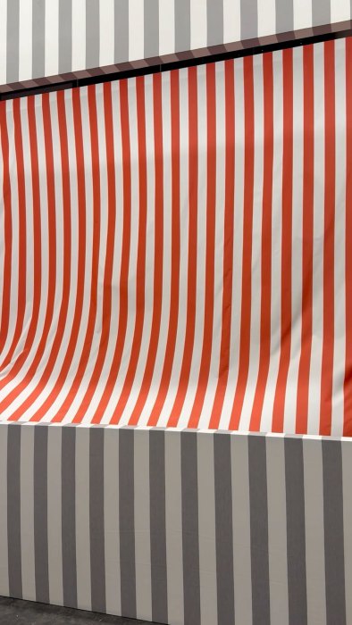A piece of red and white striped cloth (blowing by an invisible fan) was cut from a gray and white striped wall.