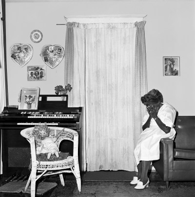 A black woman sitting on her arm on the sofa with face in her hands. Hanging on the wall of her living room, there are two heart-shaped photos.