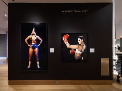 View of the museum wall painted in black. There are two colorful photos of native women hanging on it, one dressed as Wonder Woman and the other dressed as a boxer.  