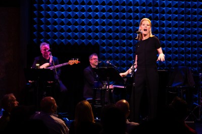 A blonde woman sings in the microphone in front of a bassist and a pianist.