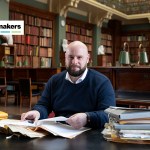 A bald man with a beard smiles at the camera in a library at a desk.