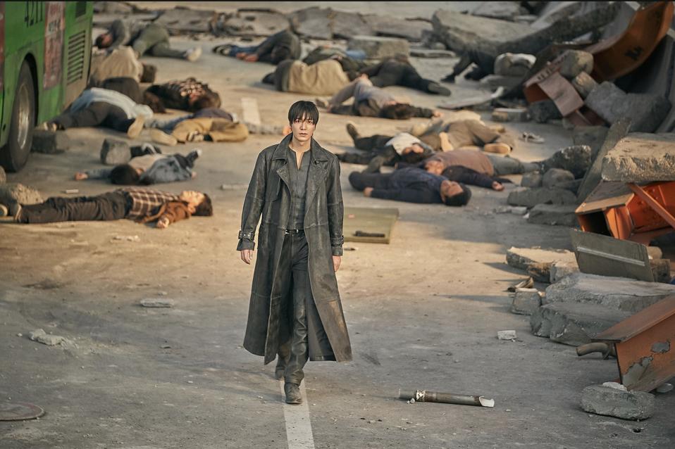 A man in long fur coat walks along the street where corpses are throwing.
