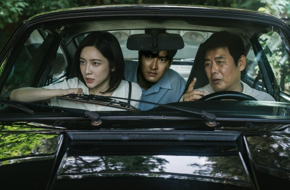 Two men and a woman are sitting in the car discussing someone.