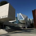 The exterior of a newly built museum displays three different structures. On the left is a rectangular shape made of shallow wood that extends out on small stairs. In the center is a silver building with angles and glass. On the right is a sculpture of Richard Serra made of brown steel.