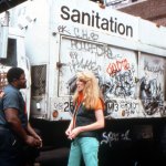 A white woman talks to a black man in a uniform of a health worker. They stood in front of a white garbage truck with the words 