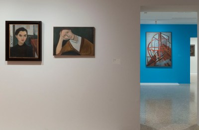 Two paintings of a woman hanging on a white wall. Visible through the portal are blue walls with abstract paintings similar to machinery hanging on them.