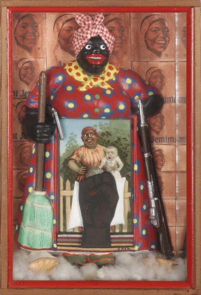 Sculpture of Aunt Jemima in the box. She held a broom in one hand and a rifle in the other. In front of her is a photo showing a mother holding a white child.