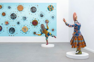 The two sculptures resemble faceless mannequins, with their arms raised in front of a large painting.