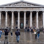 A classical structure with pillars and people walking in front of it.