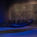 A blue hanging canoe with hands protruding out in the gallery space, a painting on the wall.