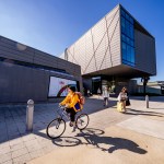 A student in a yellow coat rides a bicycle in front of the building of the Linger University Art and Design campus at a private art school in Sarasota, Florida. Ringling recently began providing AI certificates to students.