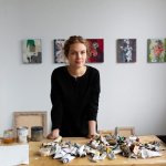 A woman stands on a table with a paint tube on it. She stood in front of five abstract paintings.
