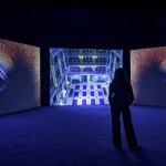Two large screens show eyes with purple iris, with a third screen showing the building in the middle. A person stands in the center of the screen.