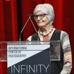 A woman stands on the podium in front of red curtains.