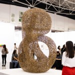 As people walk by, a sculpture stands on the pedestal of the busy art fair center.