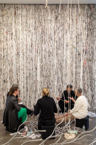 A group of people formed a circle while sitting on the sculpture. It hangs in front of them and on the wall behind them. 