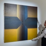 A man wearing white gloves hangs a painting on the wall. This painting shows a black cross on a yellow gradient background.