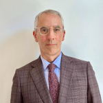 A man with glasses and gray hair is wearing a brown suit and tie.