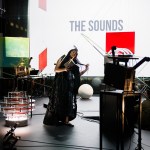A room is filled with sculptures made of film screening mechanisms, and colorful Squres hanging in the space. A long-haired black woman is holding a book and wearing a long dress because she looks like she brings a song to it.