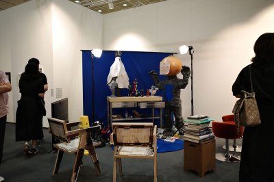 An installation consisting of a human sculpture, which is in front of a blue background and studio lights, with spherical balls and drawings. In front are chairs and books.