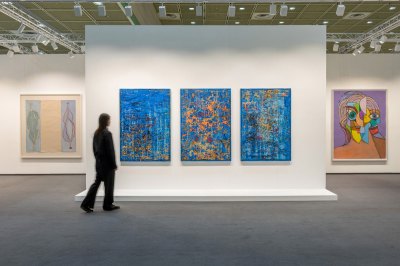 View of a person in a large art fair stall with triptychs in the center and two paintings on the walls on both sides.