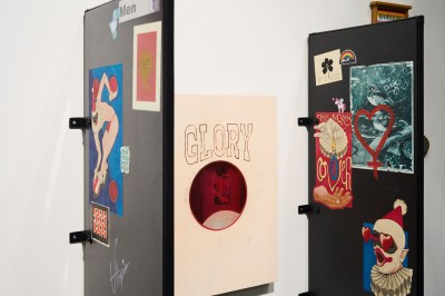 An installation resembling the dividers around a men's urinal. Where the urinal might be is a painting with the word "GLORY" and a depiction of a man receiving a blowjob.