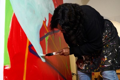 Cynthia Hawkins moves paintbrushes with light blue paint over most of the red abstract canvas. 